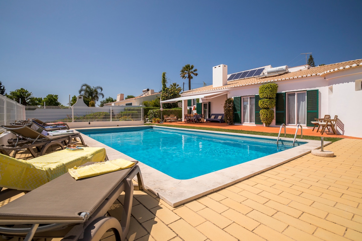 A private swimming pool is surrounded by sun loungers, with bright yellow towels draped over some chairs. A spacious patio area leads to the house, which features large sliding doors and well-maintained greenery along the exterior, under a clear blue sky.