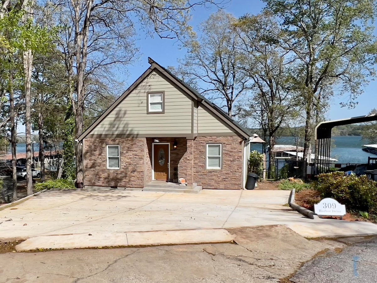 The exterior of the home showcases a combination of stone and siding, featuring a pitched roof and front door framed by windows. A spacious driveway is visible, leading to an area with nearby trees and a view of the lake in the background.