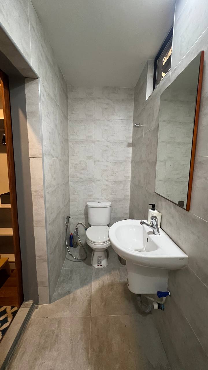 A compact bathroom is shown with modern fixtures, featuring a white toilet and a sleek sink with a mirror above. The walls are covered in light grey tiles, and a small window allows natural light to enter the space.