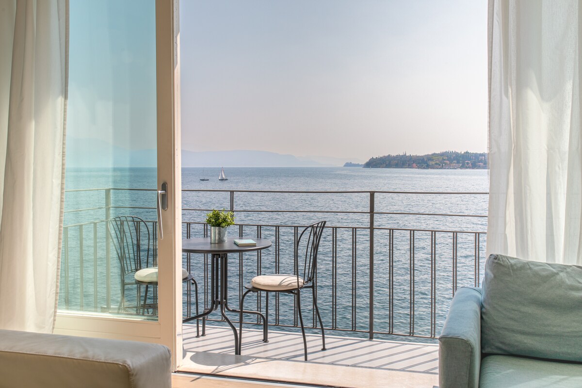 A balcony view overlooks the serene lake, accompanied by a small table and two chairs. Soft curtains frame the glass doors, allowing natural light to brighten the space. In the distance, a sailboat can be seen gliding across the water.