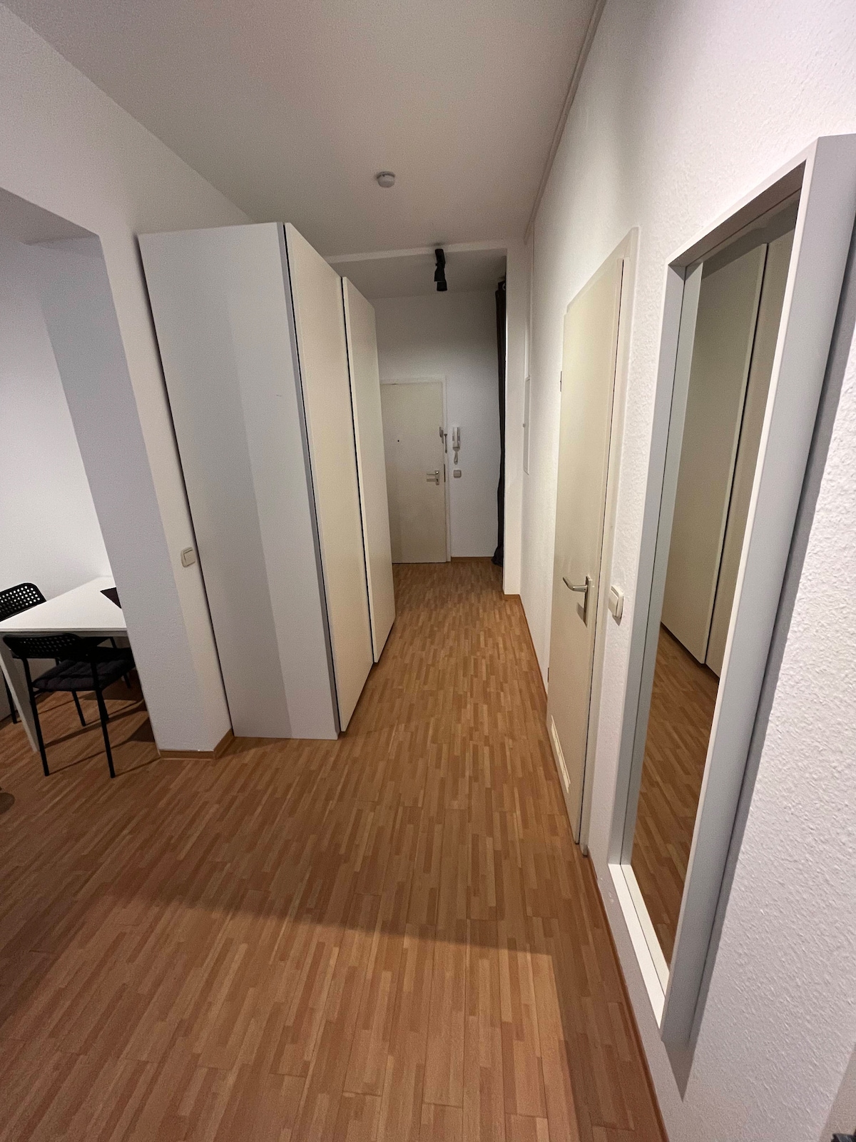 A long hallway is lined with sleek white wardrobes and a mirror. The wooden floor adds warmth to the space, while a small table with chairs is visible in the background, suggesting a functional and inviting entry area.