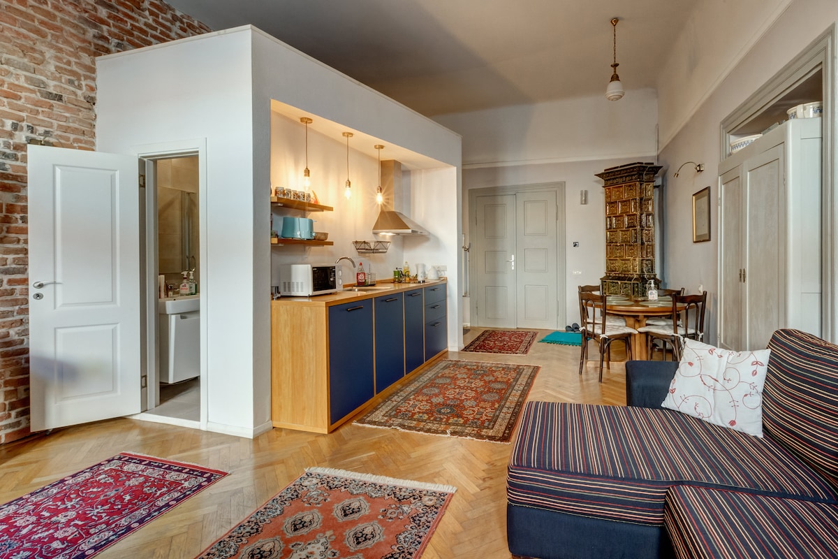The spacious living area features a blend of modern and vintage elements, including an L-shaped couch with striped fabric. A well-equipped kitchen, highlighted by wooden cabinetry and pendant lighting, is adjacent to a dining table set for four. Colorful rugs adorn the herringbone wooden floor.