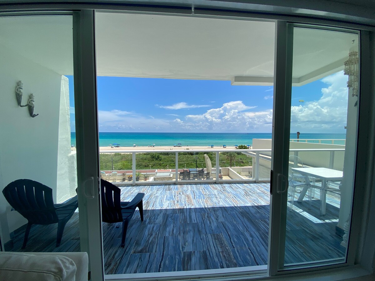 A spacious balcony features two black lounge chairs, offering a view of the beach and ocean beyond. The smooth wooden floor complements the expansive glass doors that open to the outdoor area, allowing natural light to fill the interior space. The sky is partly cloudy.