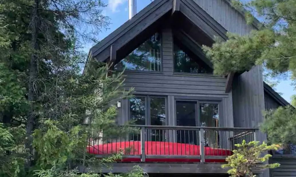 The cabin's exterior features a steep A-frame design with large glass windows, allowing natural light to illuminate the interior. A spacious deck is adorned with a striking red couch, surrounded by greenery, offering a comfortable outdoor space to relax and enjoy the views.