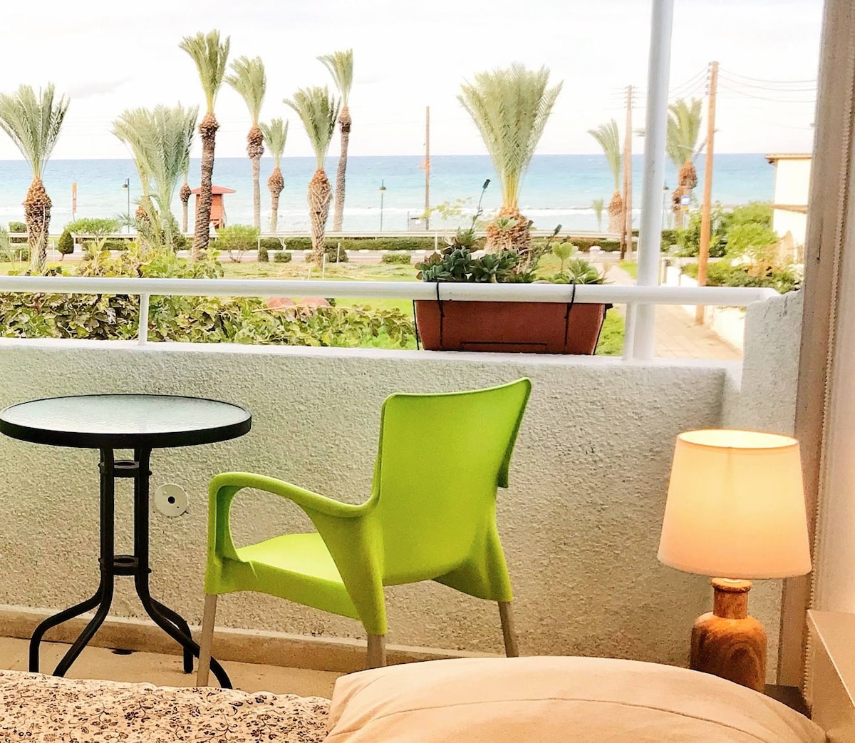 A serene balcony space features a small round table and a vibrant green chair, inviting relaxation. A lamp softly illuminates the area, while views of palm trees and the sea create a tranquil backdrop.
