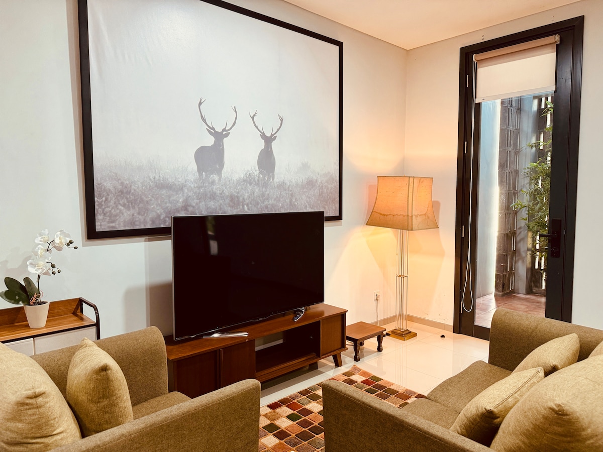 A cozy living room setup is showcased, featuring two comfortable armchairs facing a 58-inch smart TV. A large framed artwork of deer in a misty landscape is prominently displayed on the wall. A floor lamp and sliding door add warm light and access to the courtyard.