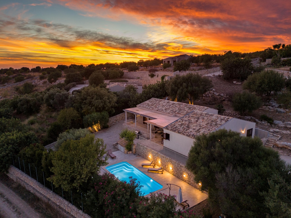 An aerial view of a villa surrounded by lush greenery and a tranquil pool. A colorful sunset lights up the sky, highlighting the villa's terrace area. Clear pathways lead to the pool, with trees and shrubs offering natural privacy around the property.