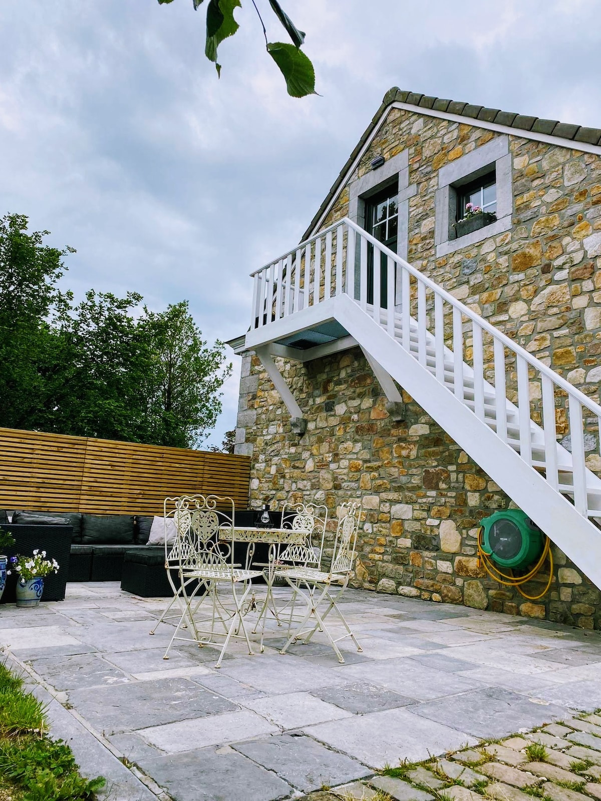 A stone structure features a white staircase leading to an upper level. A patio area is adorned with a round table and four chairs, surrounded by greenery. A seating area is visible to one side, complemented by a wooden privacy screen.