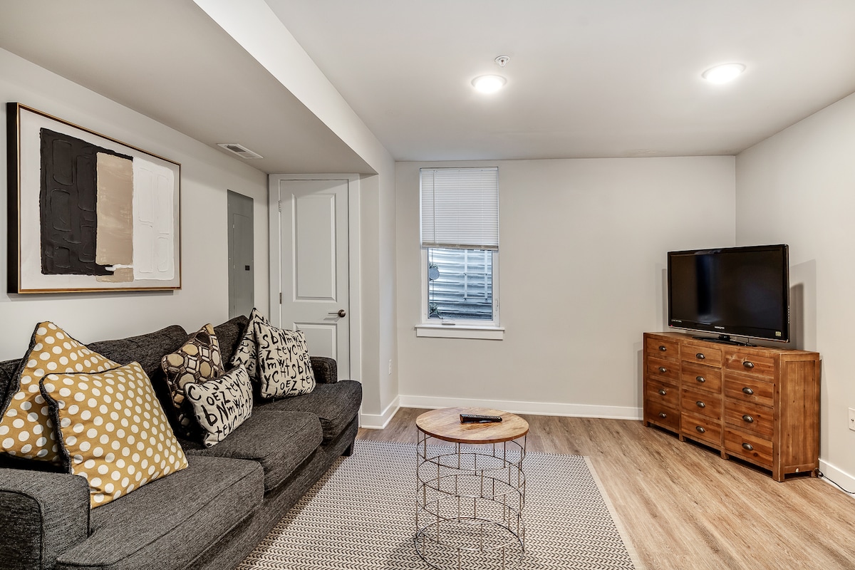 A comfortable living space is shown, featuring a gray sofa adorned with decorative cushions in various patterns. A round coffee table sits in the center on a textured area rug. A flat-screen TV is positioned against a light-colored wall, with natural light entering through a nearby window.