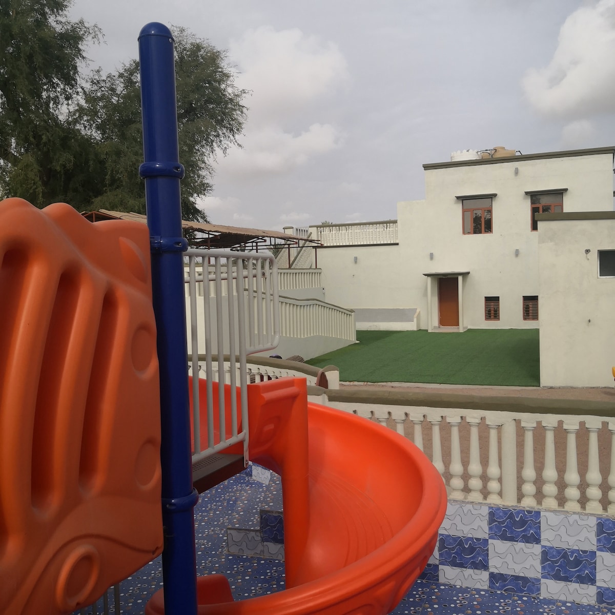 An orange slide is positioned prominently in the foreground, leading to a play area with blue tiled flooring. In the background, a green lawn is visible, with a light-colored building featuring several windows and a rooftop terrace.