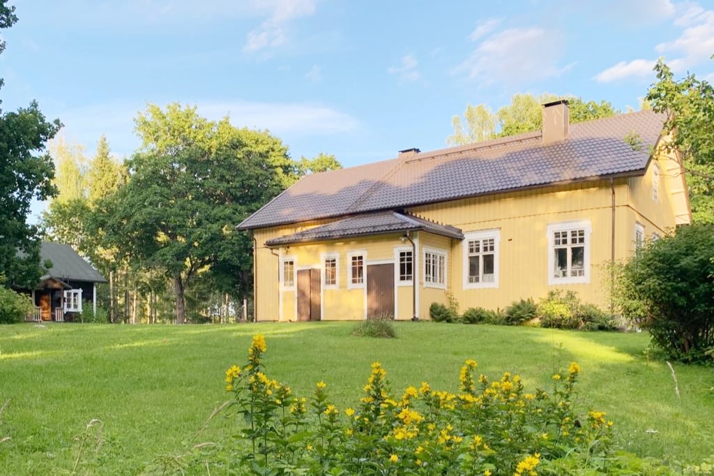 A yellow wooden house is set against a backdrop of lush greenery, featuring bright white windows and a tiled roof. A vibrant garden with yellow flowers adds color to the foreground, while additional structures can be seen in the distance.