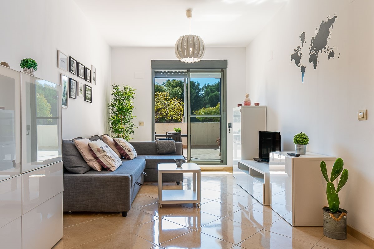 A comfortable living area is presented with a grey sofa and two armchairs. Light streams through large glass doors that open to a terrace, while a television sits on a white shelf. Decorative elements include potted plants and wall art in neutral tones.
