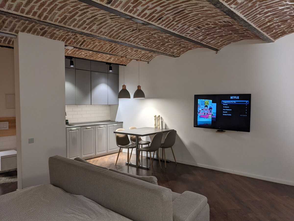 An open living space features a cozy sofa, a dining area with modern chairs, and a flat-screen TV displaying streaming options. The kitchen is equipped with sleek cabinets and provides a functional gathering area under a beautifully arched brick ceiling.
