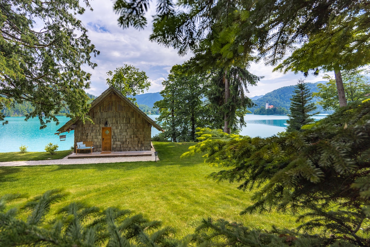 A wooden house is situated on the shore of Lake Bled, surrounded by lush greenery. The structure features a stone facade and a small patio, with expansive views of the tranquil lake and forested mountains in the background.