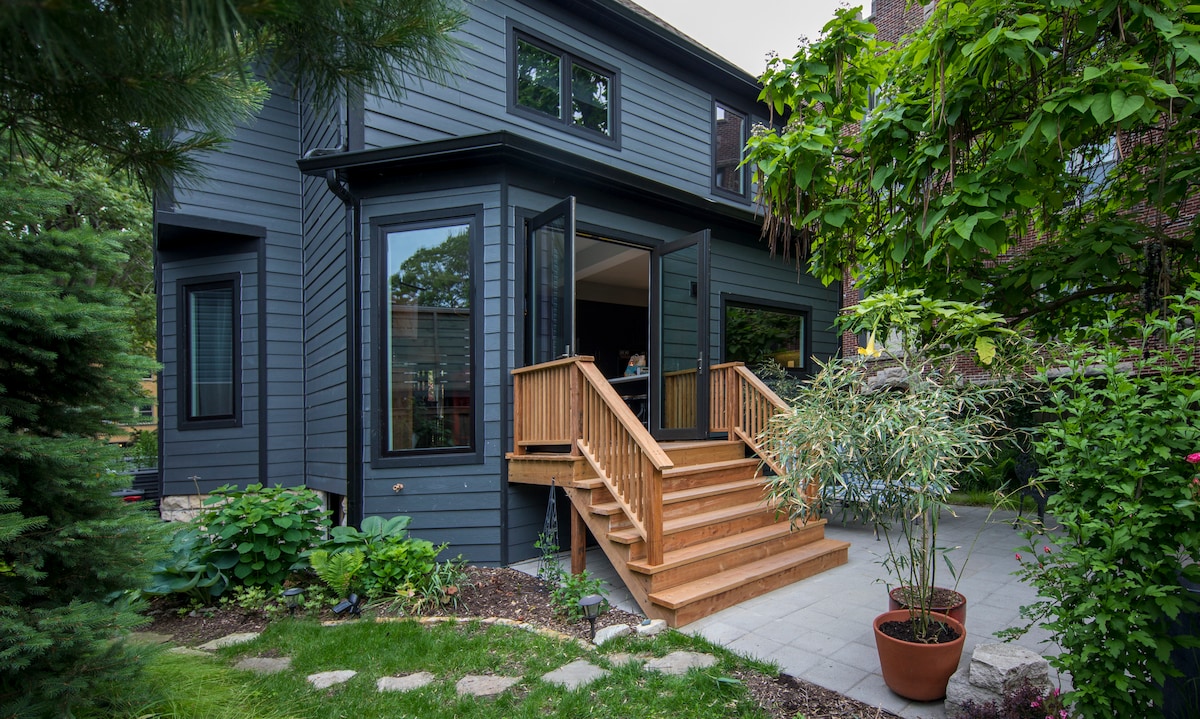 A private entrance is seen, featuring wooden steps leading to the main door. Surrounding greenery includes potted plants and lush landscaping, enhancing the inviting appearance of the entryway. Large windows provide glimpses of the interior, blending the outdoor and indoor spaces.