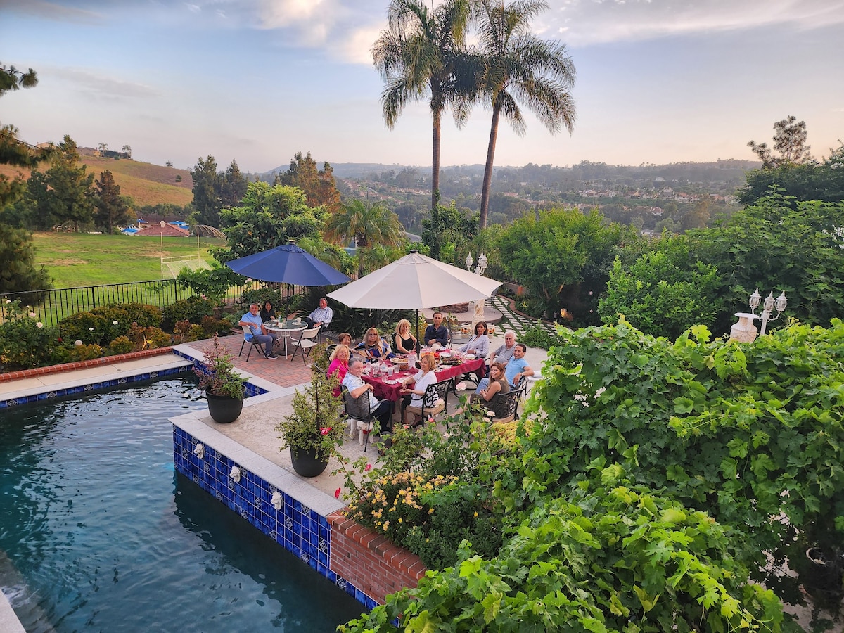 An outdoor dining area is situated beside a pool, framed by lush greenery and colorful plants. Several guests are gathered around large tables, sharing a meal under shaded umbrellas. The landscape in the background features gentle hills and greenery under a soft, evening sky.