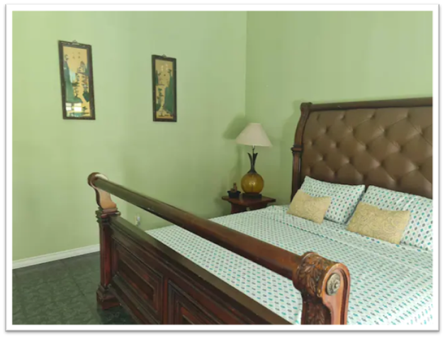 A private room features a wooden bed with a tufted headboard and soft bedding. The walls are painted in a light green hue, complemented by framed artwork. A bedside lamp adds a touch of warmth, alongside a decorative vase on the nightstand.