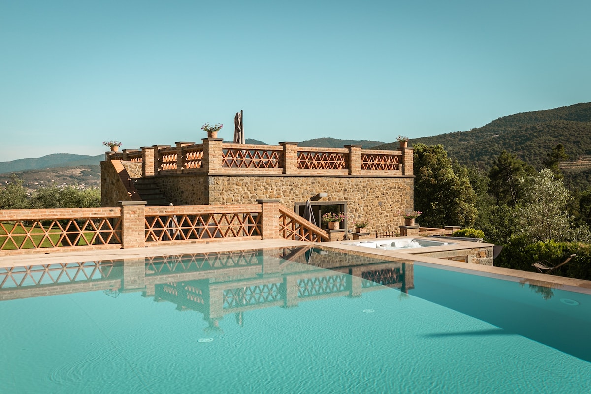 A private infinity pool reflects the clear blue sky, surrounded by a stone terrace with terracotta railings. In the background, the villa's rustic architecture is visible, framed by lush green hills and trees, creating a serene outdoor environment.
