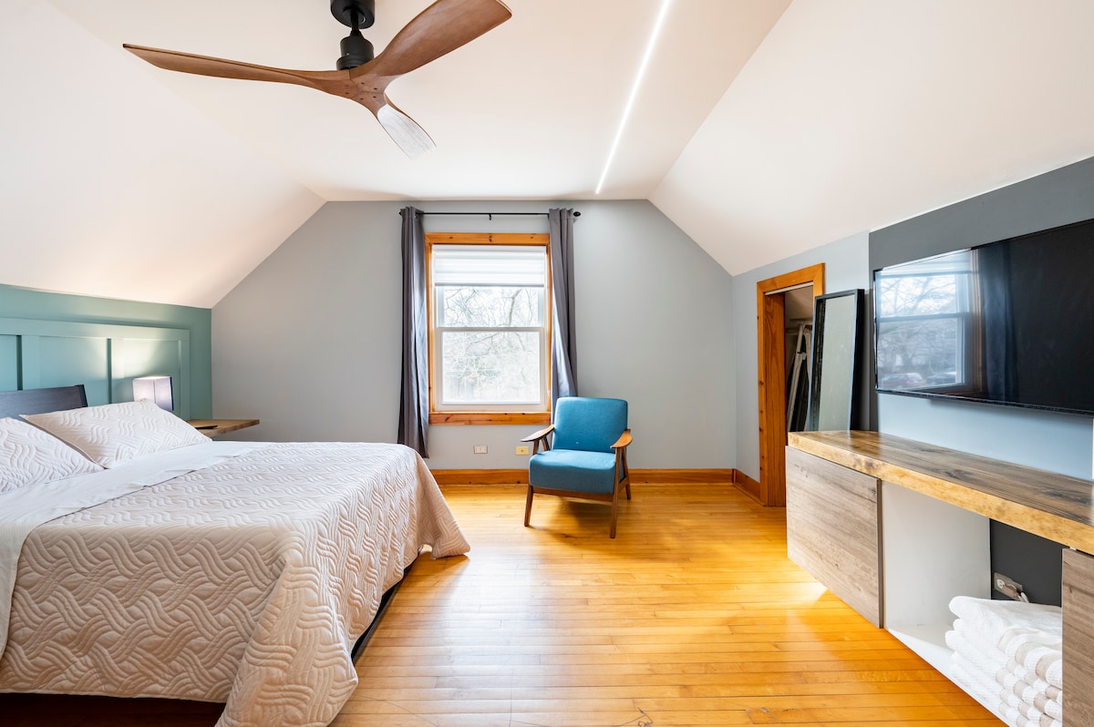 The room features a spacious bed with a textured white coverlet. A blue accent chair is positioned next to a window that allows natural light to fill the space. A wooden dresser with a television is also present, alongside warm wood flooring and neutral wall colors.