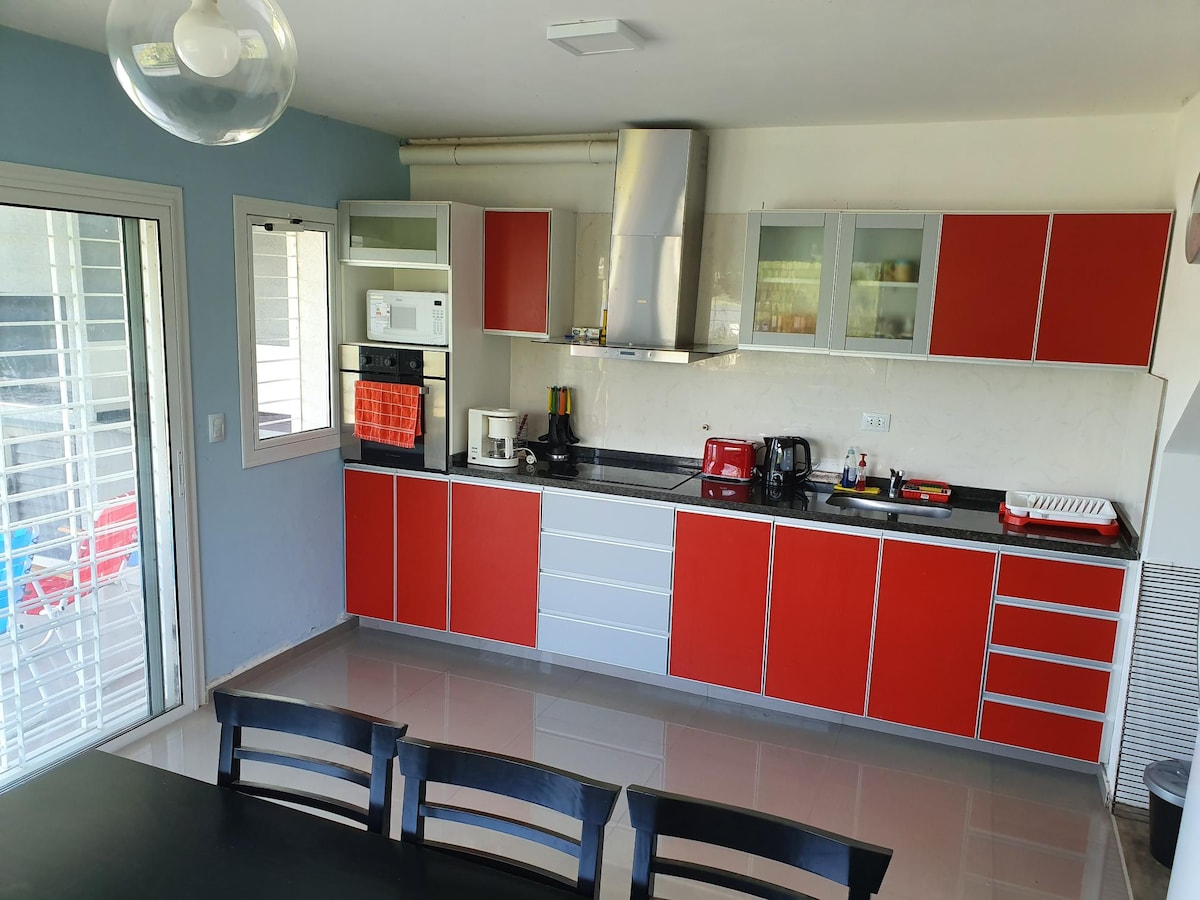A modern kitchen is featured with red and white cabinetry. Stainless steel appliances, including an exhaust hood, are present. A large window allows natural light, and a dining table with black chairs is visible in the foreground.