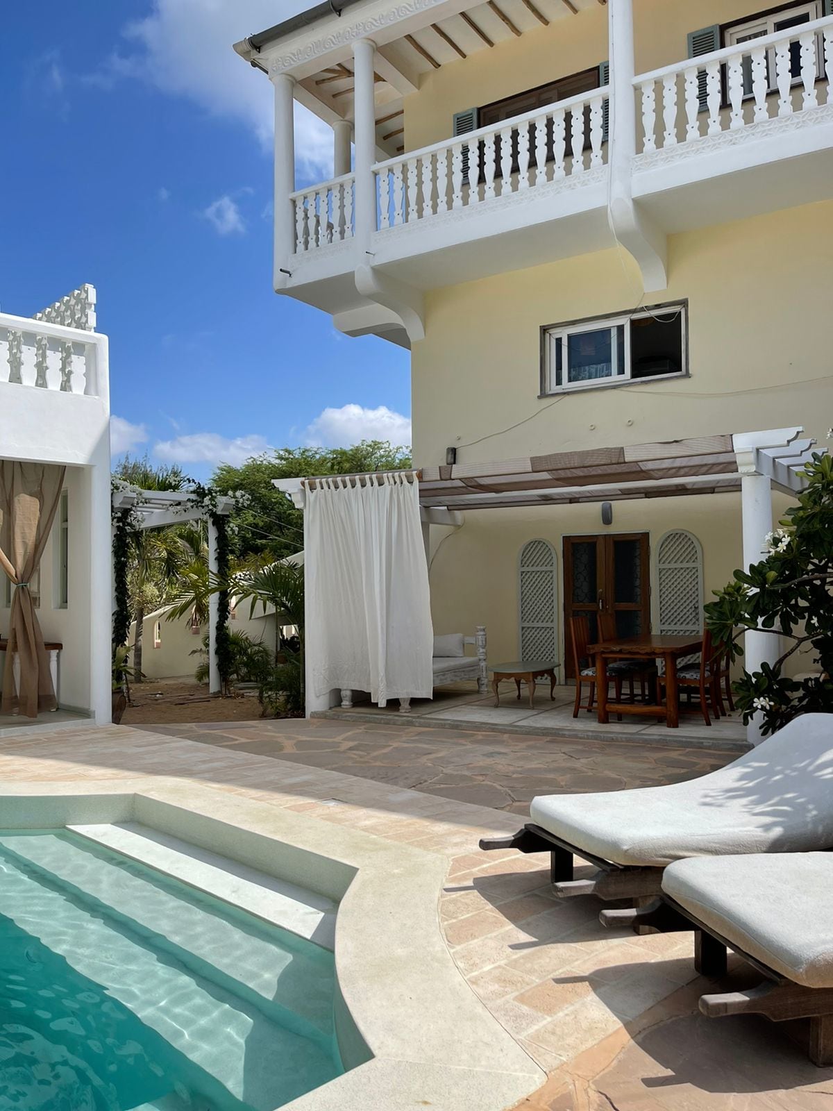 A private patio area features a swimming pool with steps leading into the water. Comfortable loungers provide space for relaxation, while a shaded dining set awaits nearby. A building with a balcony stands in the background, surrounded by tropical plants and a bright blue sky.