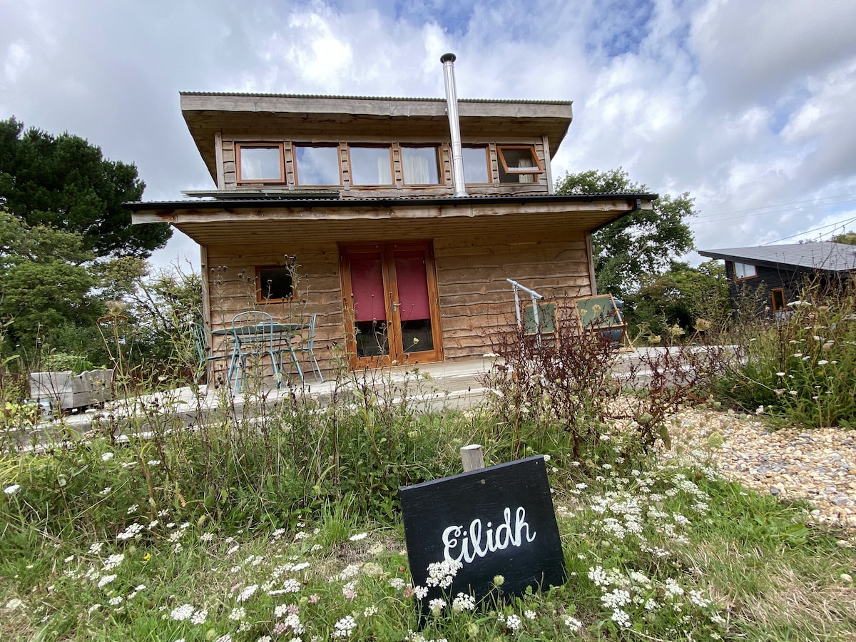 The exterior of Tiny House Eilidh is shown, featuring a wooden facade with large windows and a welcoming entrance. A small sign displays the name 'Eilidh.' Outdoor seating is visible on a patio, surrounded by natural landscaping with grass and wildflowers.