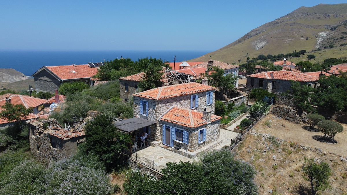 A traditional stone house with blue window frames and a terracotta roof is surrounded by lush greenery and rugged hills. The home is situated on elevated ground, providing views of the sea and nearby stone structures, highlighting the charm of the village landscape.