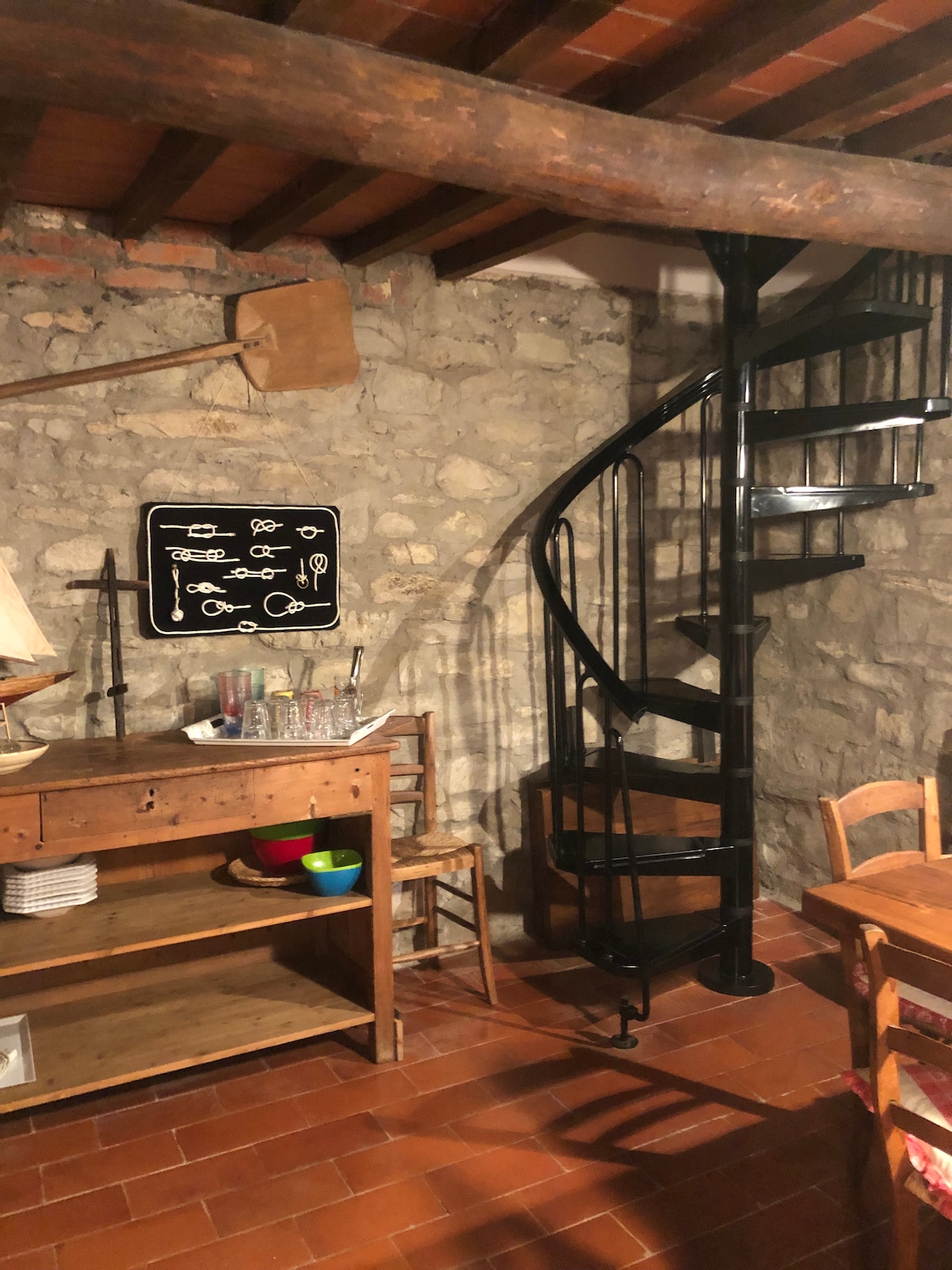 An interior space features rustic stone walls and terracotta flooring. A spiral staircase crafted from black metal ascends to the upper level. Wooden furniture, including a sideboard and several chairs, complements the warm ambiance, while kitchen utensils are illustrated on a chalkboard.