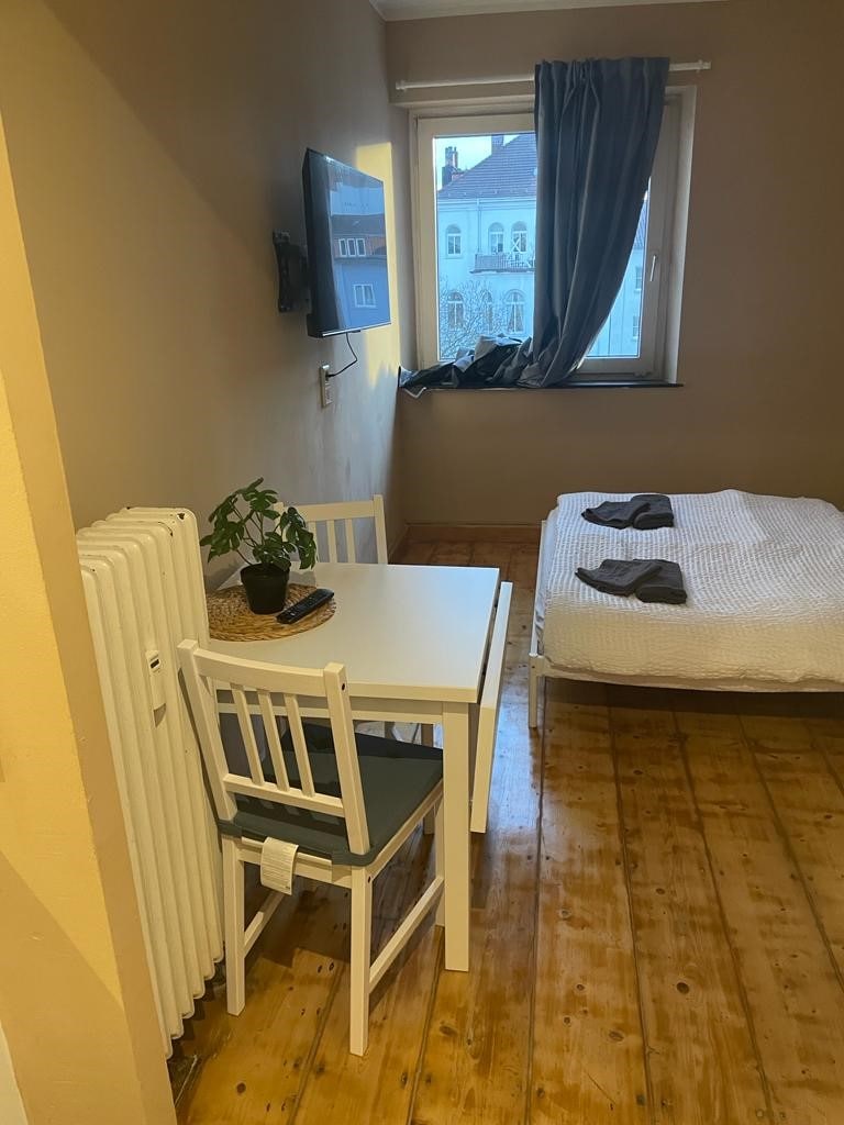 A small dining area is located beside a spacious bed, featuring a white quilt cover. A plant sits on a table, accompanied by two white chairs. Natural light enters through a window adorned with dark curtains, highlighting the warm wooden flooring.