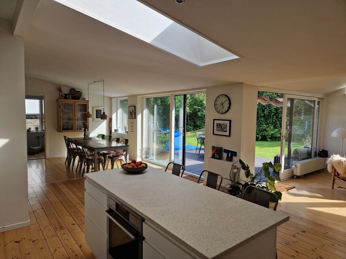 An open kitchen area showcases a spacious stone island with a bowl of fruit. Large glass doors create a seamless connection to the garden, while a dining table is visible in the background, surrounded by comfortable chairs. Natural light fills the room through a skylight.