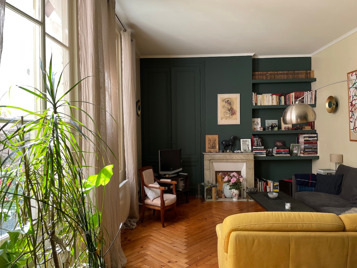 The living area features a wooden floor and a mix of contemporary and vintage decor. A leafy plant stands beside the window, adding greenery. A comfortable seating area includes an armchair and a yellow sofa, with a bookshelf and art adorning the walls.