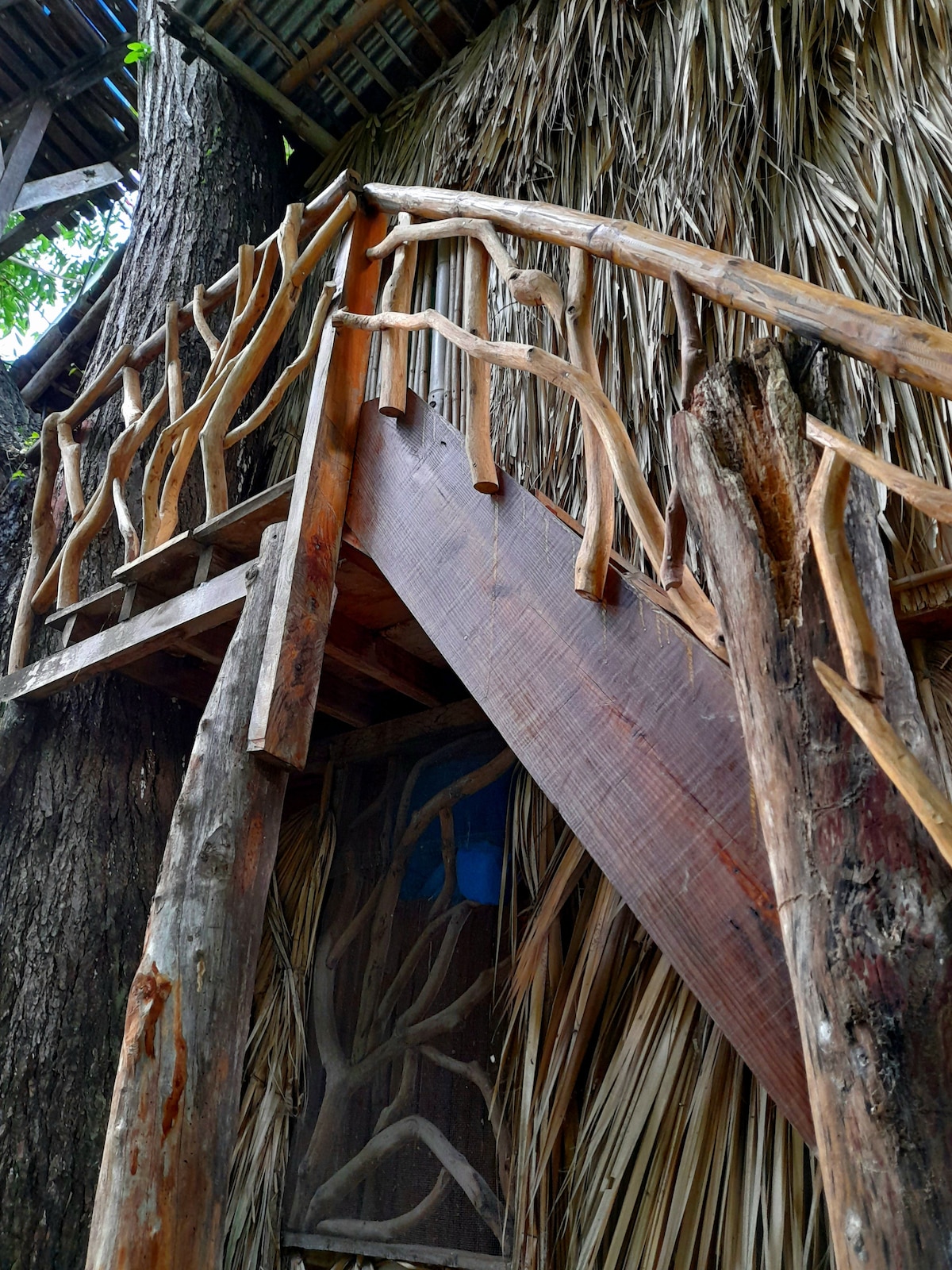 A rustic staircase leads upward, crafted from natural wood with unique, organic shapes. The railing showcases a hand-carved design that blends harmoniously with the surrounding natural materials, including thatched palm walls that create a warm and earthy aesthetic.
