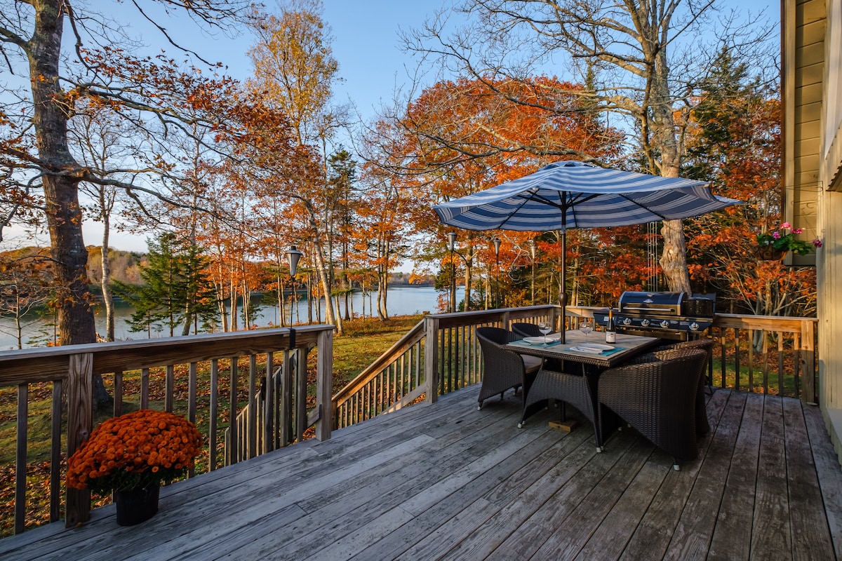 A deck area features a dining table surrounded by chairs, shaded by an umbrella. A grill is positioned nearby, with colorful flowers accentuating the space. The backdrop includes autumn foliage and a view of the water, creating a peaceful outdoor setting.