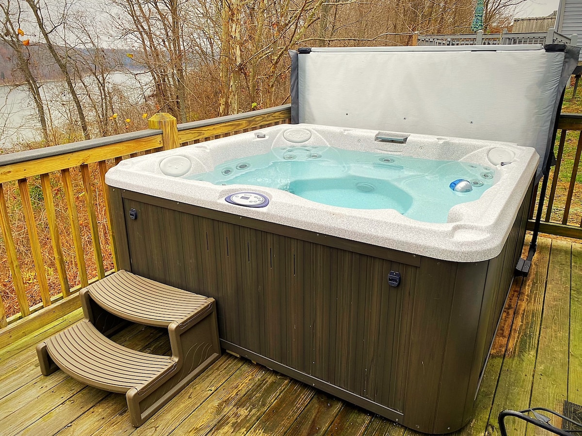 A state-of-the-art Jacuzzi J-235 hot tub is positioned on a private deck, featuring water jets and comfortable seating. Steps lead up to the tub, and the tranquil landscape of trees and a lake is visible in the background, enhancing the serene outdoor setting.