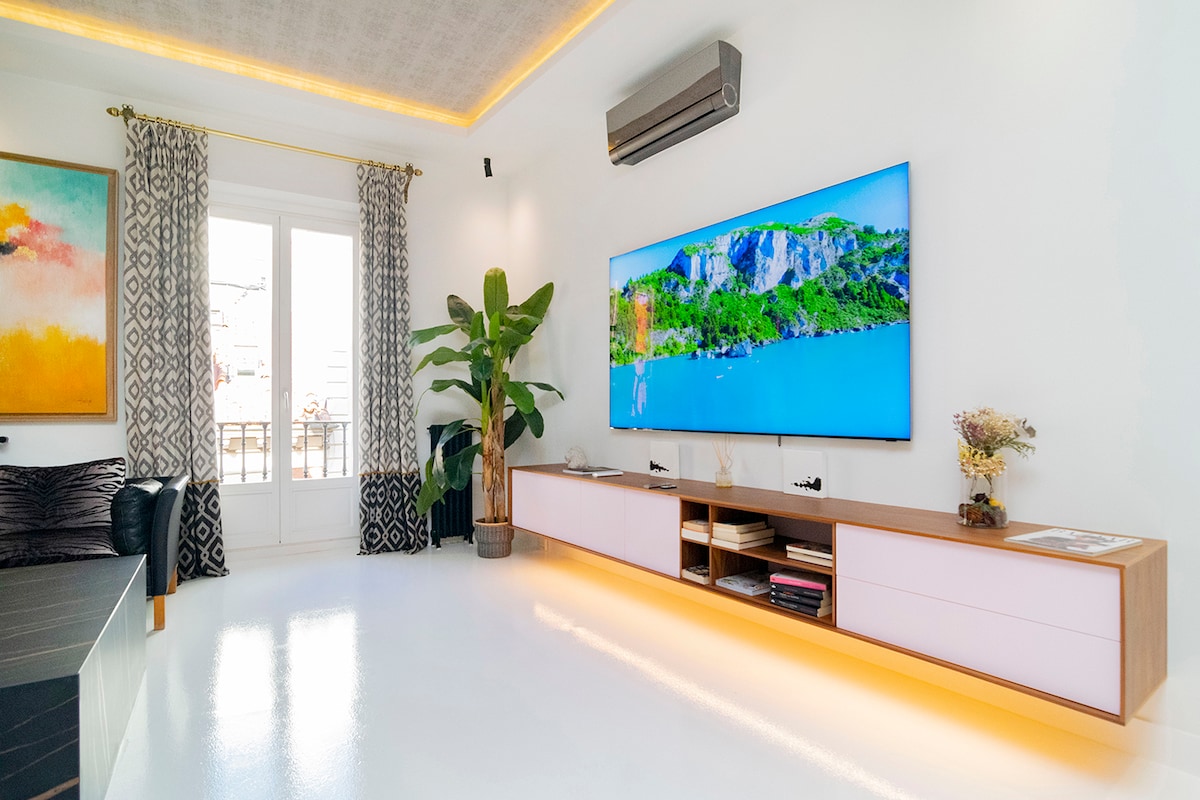 A bright living room features a large mounted TV on a minimalist console, complemented by elegant wooden drawers. A stylish black sofa is positioned nearby, with a decorative plant adding a touch of nature. Natural light fills the space through a balcony door adorned with curtains.