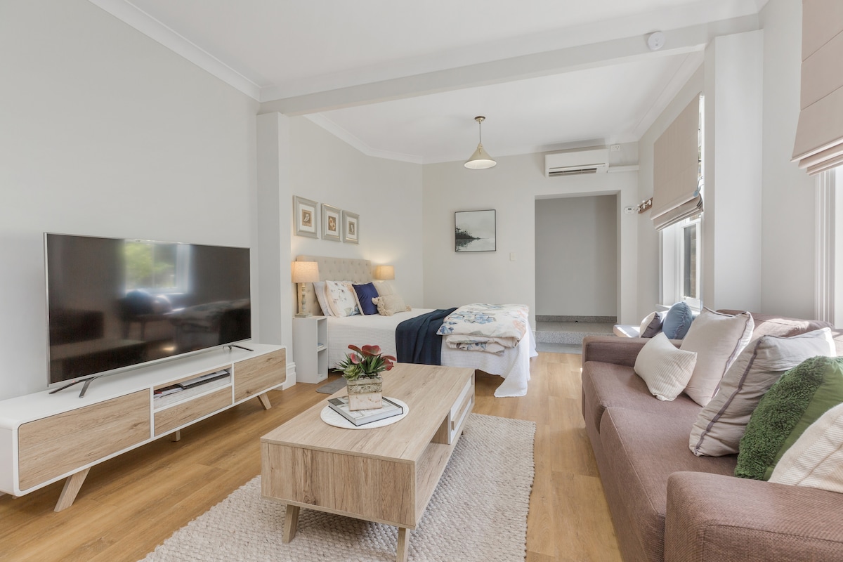 A modern living space showcases a comfortable brown sofa contrasted by a light wooden coffee table. A spacious bed with decorative pillows is positioned against a light wall, and a large TV is mounted for viewing. Soft natural light filters through the windows.