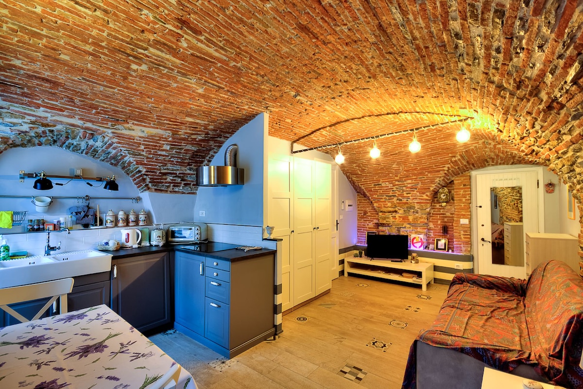 A renovated studio features exposed brick arches and soft lighting, creating a cozy atmosphere. The kitchen area is equipped with modern appliances, while the living space includes a comfortable sofa bed and a television. A dining table is positioned near the kitchen, enhancing usability.