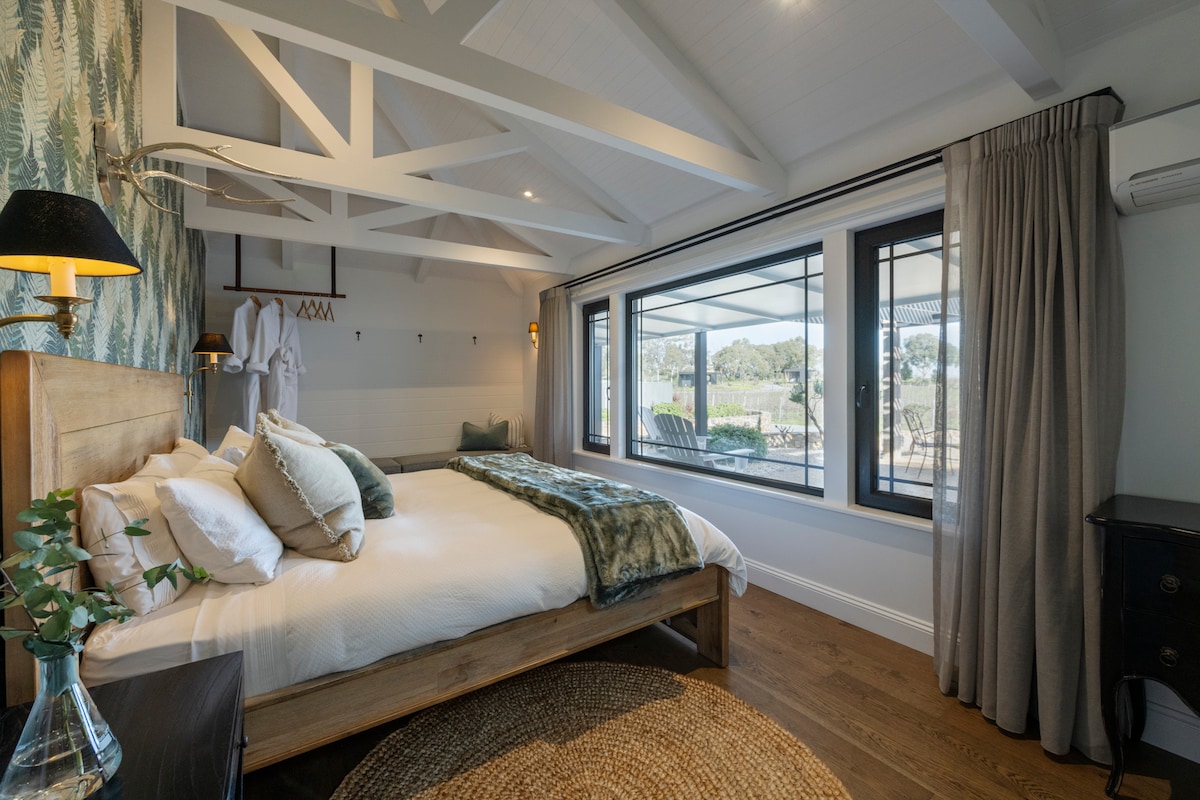 A spacious bedroom features a king bed with luxury linens and a decorative throw. Natural light filters through large windows, showcasing views of the vineyard. Elegant decor includes a wooden bed frame, bedside lamp, and soft area rug, creating a serene atmosphere.