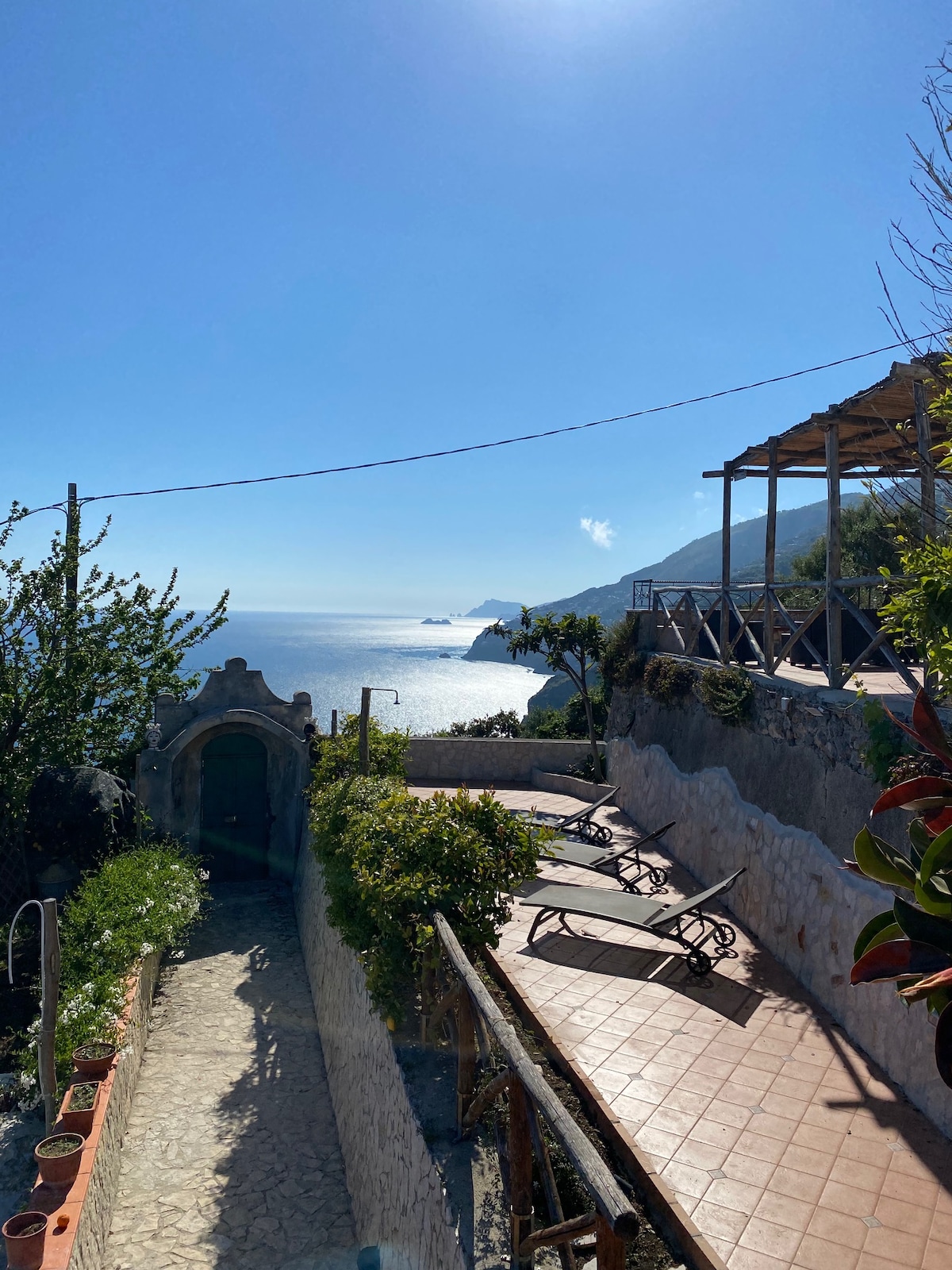A scenic path leads to an outdoor terrace with sun loungers, overlooking the sparkling sea. Lush greenery lines the walkway, and distant cliffs are visible against a clear blue sky. The tranquil atmosphere is enhanced by the distant sounds of nature.