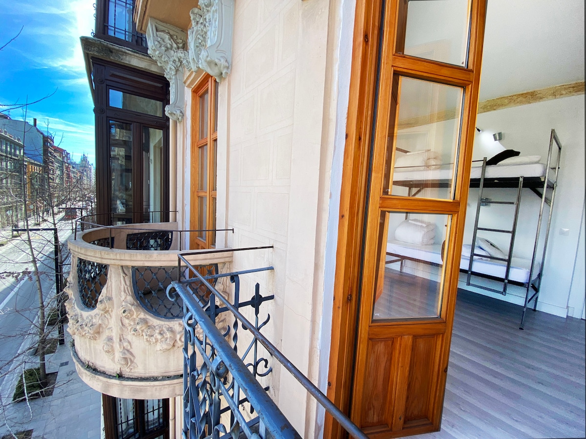 A balcony with ornate wooden details overlooks a quiet street, offering a view of the building's façade. Inside, a room is visible, featuring a bunk bed and ample space. Natural light fills the area, highlighting the wooden floors and architectural elements.