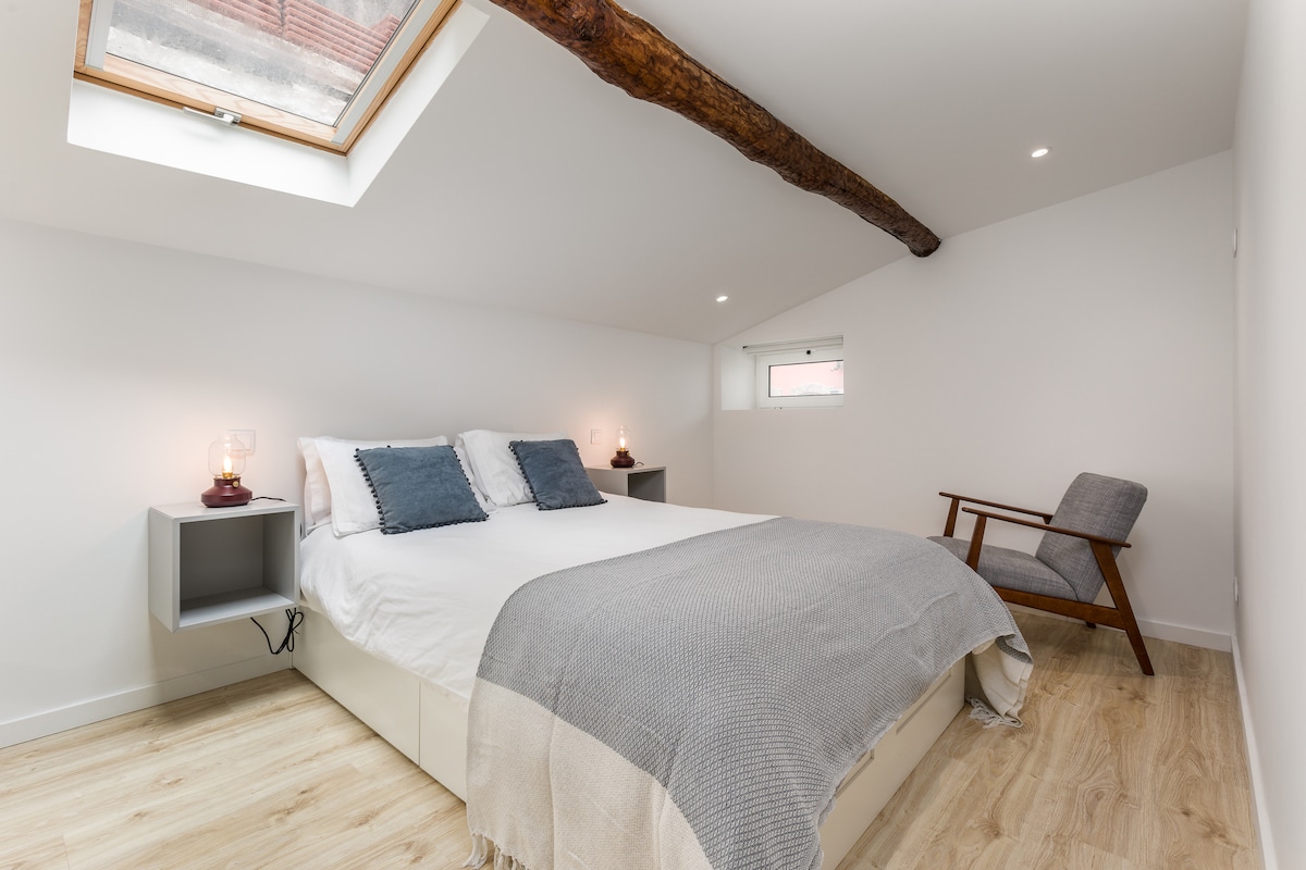 A spacious sleeping area is highlighted by a large window with a wooden beam overhead. A plush bed is flanked by nightstands with lamps, while a comfortable chair is positioned nearby. Soft neutral tones create a calm and restful environment.