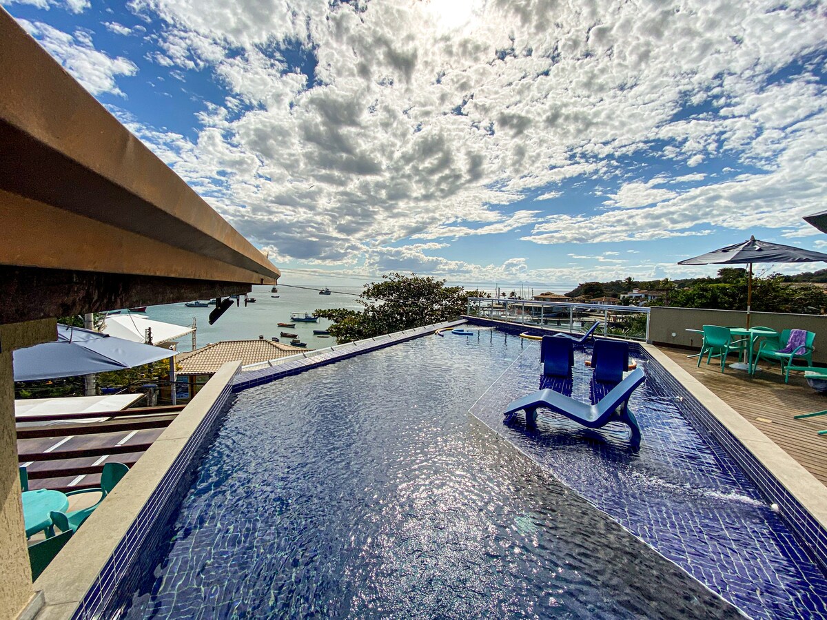 A shimmering infinity pool overlooks the coastline, offering views of the water and surrounding scenery. Sun loungers are positioned beside the pool, complemented by umbrellas providing shade. The sky showcases a blend of clouds and sunlight, enhancing the tranquil atmosphere.
