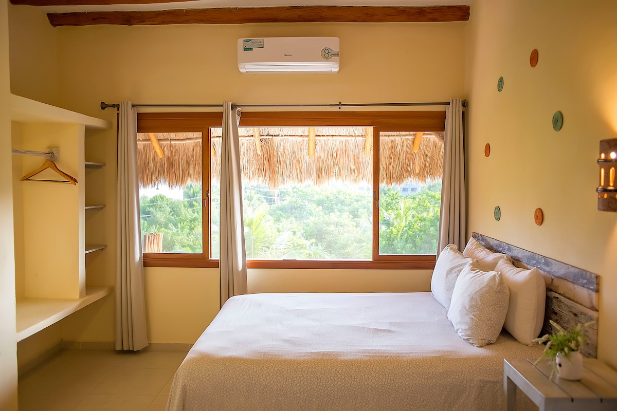 A spacious sleeping area is presented with a comfortable bed adorned with crisp linens. The room features large windows with straw-covered frames, allowing natural light to fill the space while offering views of surrounding greenery. Simple shelves and a hanging area are included for convenience.