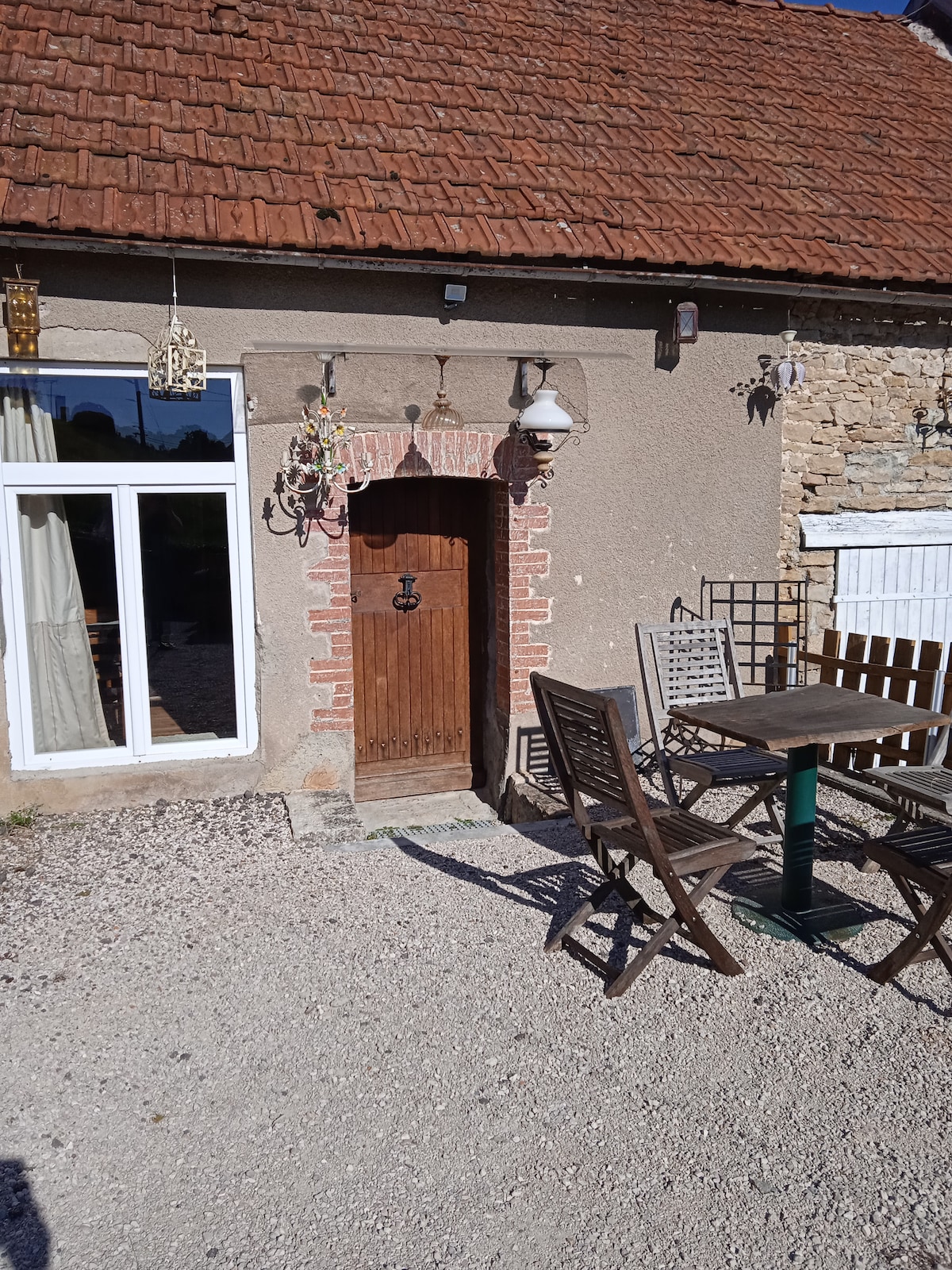 The entrance to a cozy home is framed by a rustic wooden door. A set of wooden patio furniture is arranged outside, inviting relaxation. Natural light streams through large windows, illuminating the gravel pathway and textured stone façade.