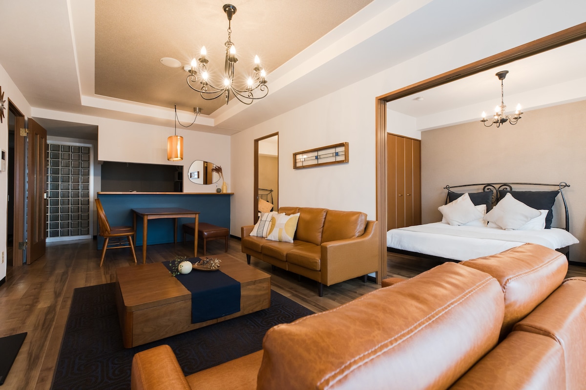 A spacious living area features a leather sofa and coffee table, with a dining space highlighted by a pendant light. A well-appointed kitchen area is visible in the background. Beyond, a bedroom with a large bed is partially visible, enhancing the room's overall comfort.