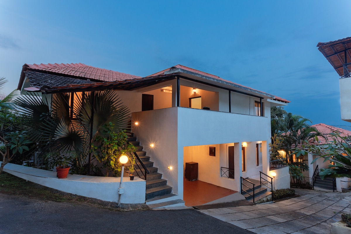 A two-story villa is shown, featuring a combination of white and terracotta exteriors. Several potted plants decorate the entryway, and soft lighting accents the staircase, guiding visitors to the upper level. A pathway leads through a landscaped area, embraced by palm trees.