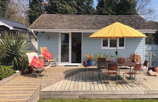 A self-contained unit is showcased with a light blue exterior, featuring a spacious wooden deck area. A large yellow umbrella shades a dining table surrounded by multiple chairs. Lounge chairs are positioned nearby, complemented by vibrant cushions and potted plants, enhancing the outdoor setting.