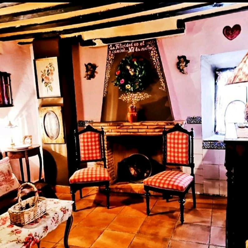 A cozy seating area is presented with two checker-patterned chairs positioned near a rustic fireplace. A flower arrangement decorates the mantle, complemented by soft lighting from a nearby table lamp. The walls feature charming decor, enhancing the warm ambiance of the space.