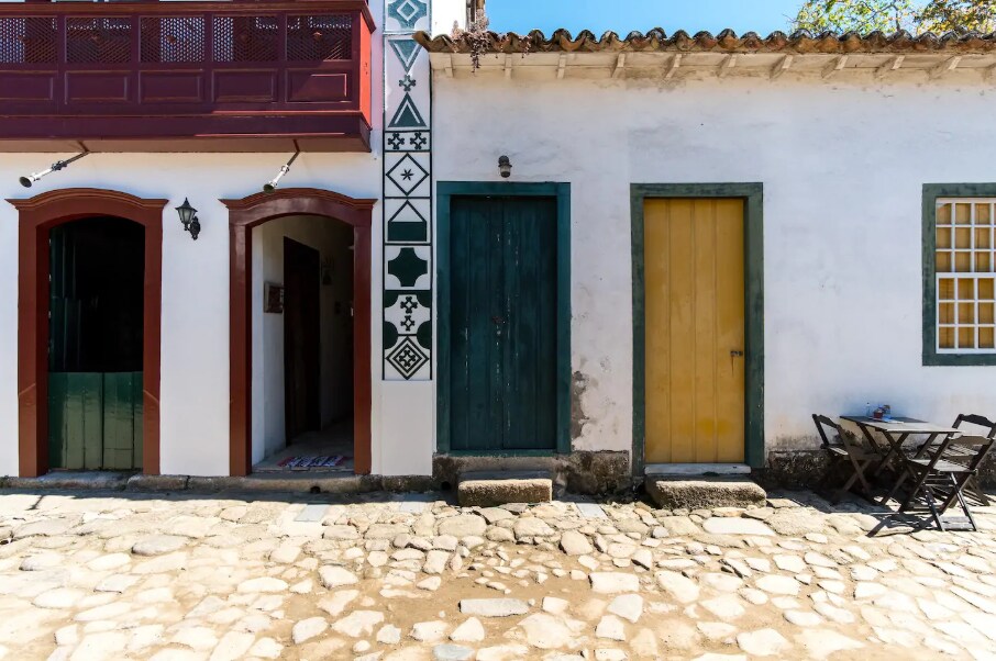 A charming street view displays rustic colonial architecture with vibrant colors. Distinctive geometric patterns decorate the wall beside a double doorway. A green door and a bright yellow door lead into separate spaces. A small table and chairs are positioned on the cobblestone pathway.