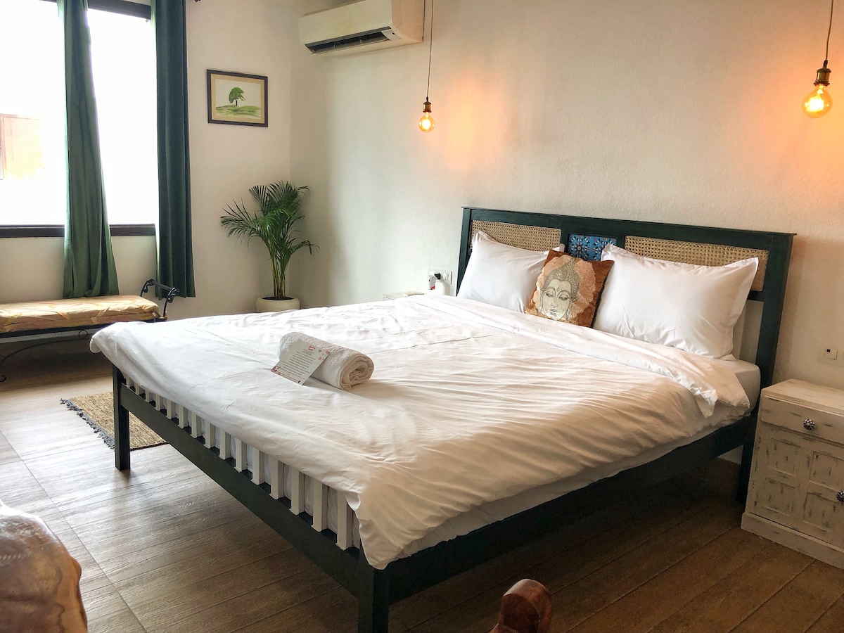 A spacious bedroom is equipped with a large bed featuring crisp white linens and decorative pillows. Soft lighting is provided by hanging bulbs, while natural light filters through green curtains. A small seating area is visible by the window, accompanied by a plant.